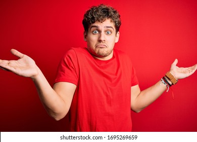 Young blond handsome man with curly hair wearing casual t-shirt over red background clueless and confused expression with arms and hands raised. Doubt concept. - Powered by Shutterstock