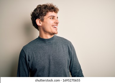 Young Blond Handsome Man With Curly Hair Wearing Casual Sweater Over White Background Looking Away To Side With Smile On Face, Natural Expression. Laughing Confident.