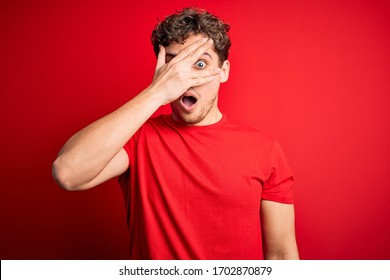 Young blond handsome man with curly hair wearing casual t-shirt over red background peeking in shock covering face and eyes with hand, looking through fingers with embarrassed expression.