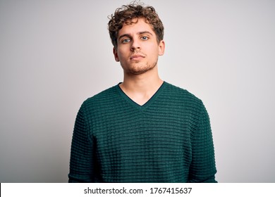 Young blond handsome man with curly hair wearing green sweater over white background Relaxed with serious expression on face. Simple and natural looking at the camera.