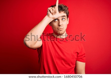 Young blond handsome man with curly hair wearing casual t-shirt over red background making fun of people with fingers on forehead doing loser gesture mocking and insulting.