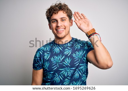 Young blond handsome man on vacation with curly hair wearing casual summer t-shirt Waiving saying hello happy and smiling, friendly welcome gesture
