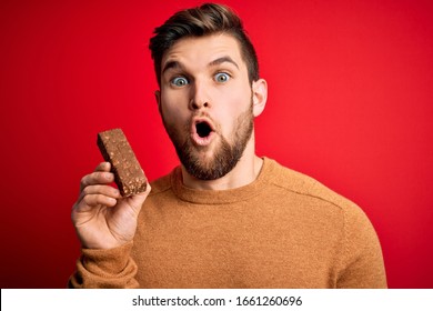 Young blond man with beard and blue eyes eating healthy chocolate protein bar scared in shock with a surprise face, afraid and excited with fear expression - Powered by Shutterstock