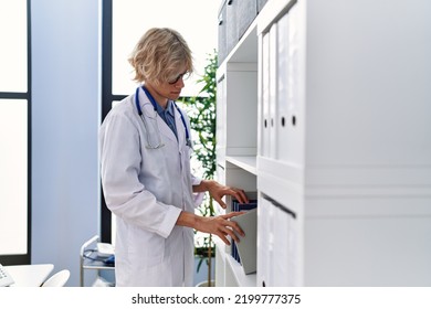 Young Blond Man Doctor Holding Book On Shelving At Clinic