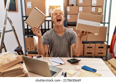 Young blond man holding packages working at online shop angry and mad screaming frustrated and furious, shouting with anger looking up. 