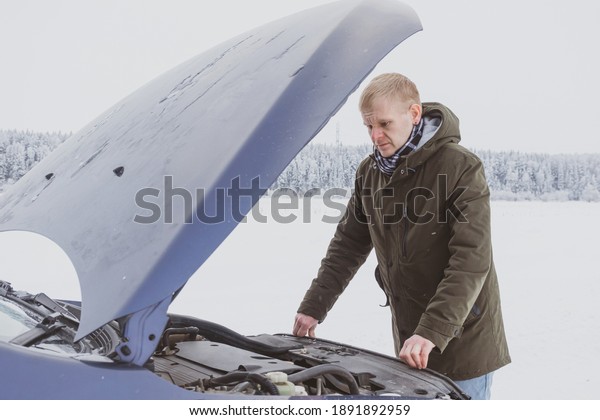 A young blond man in a jacket\
looks under the hood of a car at the engine. Malfunctions on the\
winter track, car breakdown. The driver is confused and\
upset.