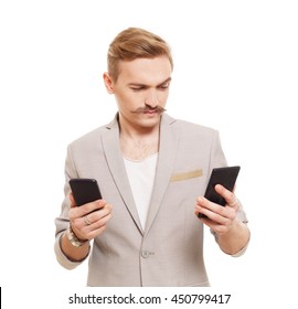 Young Blond Man With Mustache Choose Between Two Cell Phones. Guy Looks At Mobile, Selecting Which To Buy. Comparing Smartphones In Hands. Male Portrait Isolated At White Background