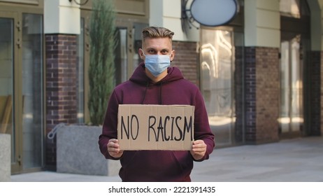 A Young And Blond Man Protester Activist Is Wearing A Medical Mask Holds A Cardboard Poster With The Slogan NO RACISM. A Protest Against Racism And Violence During Quarantine. Stop Racism Concept.