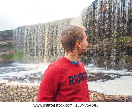 A young blond man in a red sweater looks at the Waterfall on a gloomy cloudy day, Estonia