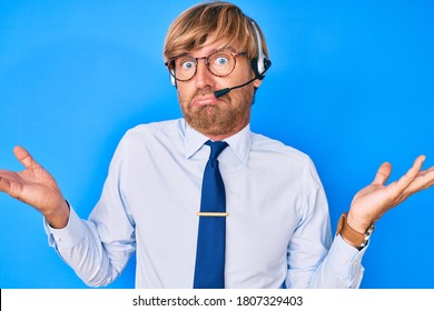 Young Blond Man Wearing Call Center Agent Headset Clueless And Confused With Open Arms, No Idea And Doubtful Face. 