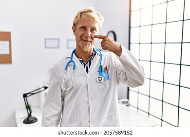 Young Blond Man Wearing Doctor Uniform And Stethoscope At Clinic Pointing With Hand Finger To Face And Nose, Smiling Cheerful. Beauty Concept 