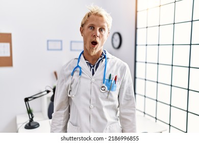 Young blond man wearing doctor uniform and stethoscope at clinic in shock face, looking skeptical and sarcastic, surprised with open mouth 