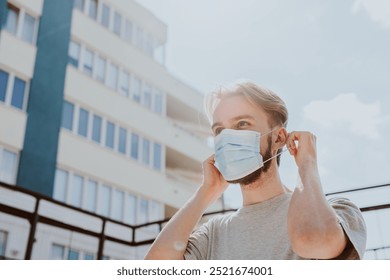 Young blond man wearing a mask outdoors. The lond man adjusts his mask in front of a building, city background. Mask-wearing is essential for safety.The man ensures his mask fits properly on his face. - Powered by Shutterstock