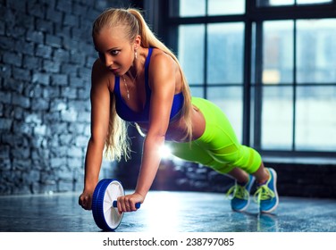 Young blond slim woman do abdominal exercises with wheel in modern interior on window background.