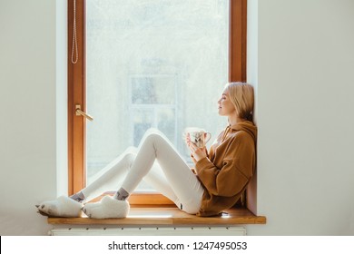 Young Blond Teen Woman In Funny Fur Slippers Relaxing With Hot Coffee Or Tea On Window Sill In Empty Cozy Home In Sunny Winter Day. Side View