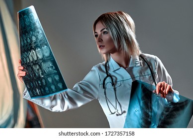 A young blond woman doctor dressed in a white uniform holds an MRI of the brain isolated on a dark gray background.