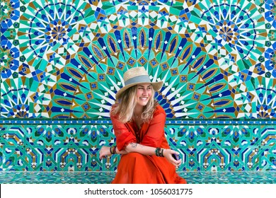Young blond woman in hat and in red dress is sitting on a multicolored mosaic wall background and smiling happily.