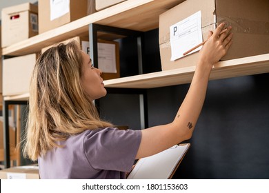 Young blond woman with list of orders and pencil checking address of receiver on packed box while standing by shelf in storage room