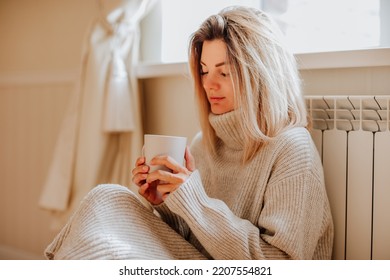 Young blond woman in long winter beige sweater is holding a cup of coffee and posing at home near the radiator. Winter season concept. Focus is at hands. Economy program