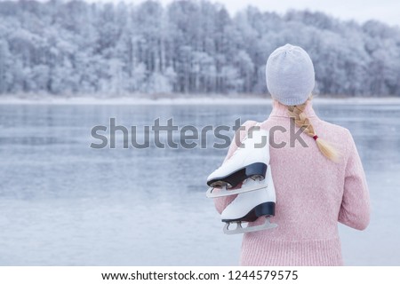 Young blond woman in pink sweater and hat staring at ice of lake and holding white skates over shoulder in freezing winter day. Back view of ice skater. Outdoor activities on weekends in cold weather.