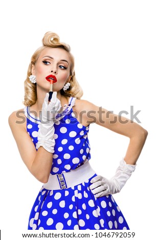 Young blond woman in pin-up style blue dress in polka dot, applying lipstick, isolated over white background. Caucasian model posing in retro fashion and vintage concept.