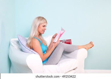 Young Blond Woman Sat In An Armchair Opening Her Birthday Card.