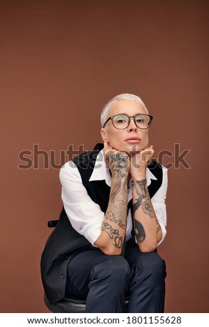 Young blond woman with short hair and arms covered with tattoos sitting on chair in front of camera while keeping hands under chin in isolation