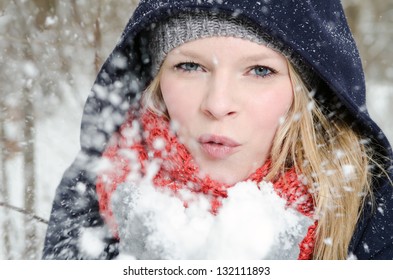 Young Blond Woman Smiles With Beanie And Scarf Blows In A Handful Of Snow