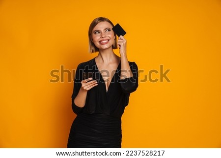 Young blond woman smiling while posing with cellphone and credit card isolated over yellow background