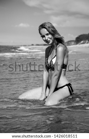young blond woman in swimsuit at sea surf with foam sitting under sun rays, looking at camera, smiling, monochrome