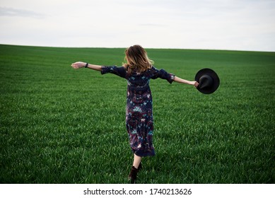 Young Blond Woman, Wearing Long Dark Boho Dress, Holding Black Hat Running Away In Green Field In Spring. Model Posing Outside In Meadow. Hippie Musician At Natural Environment.