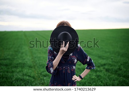 Young blond woman, wearing long dark boho dress, holding black hat covering face on green field in spring. Model posing outside in meadow. Hippie musician at natural environment.