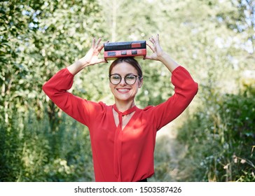 Young blond woman, wearing red shirt and eyeglasses, holding two books on her head. Student with black and red books, smiling. Studying fun. Pretty girl, balancing with books on her head, 