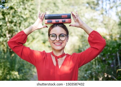 Young blond woman, wearing red shirt and eyeglasses, holding two books on her head. Student with black and red books, smiling. Studying fun. Pretty girl, balancing with books on her head, 