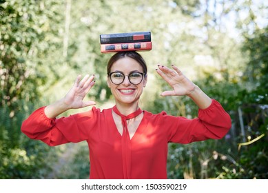 Young blond woman, wearing red shirt and eyeglasses, holding two books on her head. Student with black and red books, smiling. Studying fun. Pretty girl, balancing with books on her head,