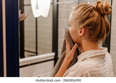 Young Blond Woman Wiping Face With Towel In Bathroom In Front Of Mirror