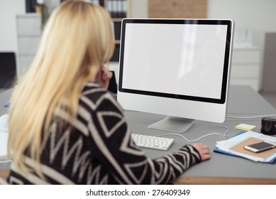 Young Blond Woman Working On A Desktop Computer At Her Desk In The Office, Viewed From Behind With A Blank White Screen