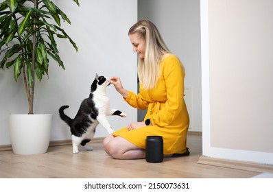 Young Blond Woman With Yellow Dress Kneeling On The Floor  Feeding Snacks To Her Cat Having The Treat Jar Standing Right Next To Her