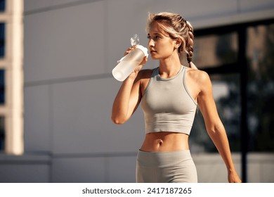 Young blonde athlete girl drinking water from sports bottle during morning jogging in the city. Female fitness model monitors the body water balance to avoid dehydration. Healthy lifestyle. - Powered by Shutterstock