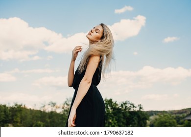 Young blonde attractive woman with long hair, wearing black maxi dress, outdoors, standing in a field in wind, on blue sky background.