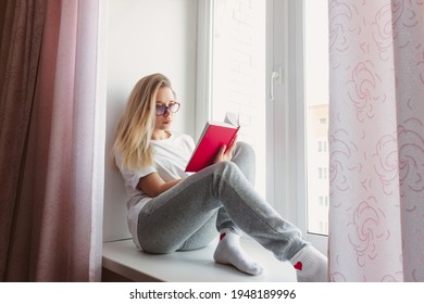 A young blonde beautiful woman Introvert in the white t-shirt and gray sweatpants with glasses is sitting on the windowsill and reading a book with a red cover. Self-isolation. Lock down