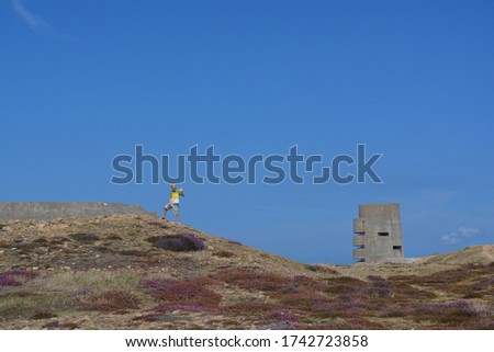 Young blonde boy stood in the sunshine on the near horizon against a clear blue sky in the wild rocky countryside with purple heather. A World War 2 concrete bunker fortress looms in the background.