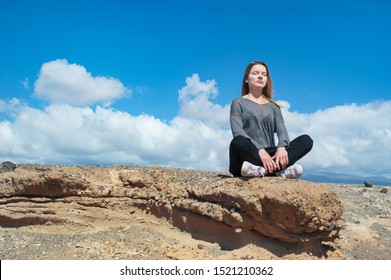 Young Blonde Caucasian Female Dressed In A Neutral Outfit, Sitting In A Relaxing, Meditative Position On A Rocky Ground, With A Calm Look On Her Face And Her Eyes Closed, In Tenerife, Canary Islands