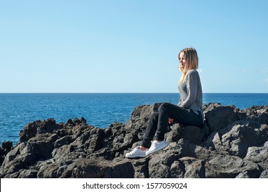 Young Blonde Caucasian Female Dressed In A Neutral Outfit, Sitting In A Relaxing, Meditative Position With A Nostalgic Look On Her Face, With Her Body Turned Towards The Ocean And The Rocky Coast