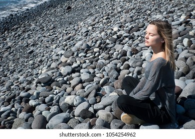 Young Blonde Caucasian Female Sitting In A Relaxing, Meditating Position With Eyes Closed, Wearing A Neutral Outfit On The Beach, In Harmony With The Surroundings, Under The Bright Morning Sunlight 