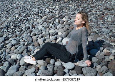 Young Blonde Caucasian Female Sitting In A Relaxing, Meditating Position With Eyes Closed, Wearing A Neutral Outfit On The Beach, In Harmony With The Surroundings, Under The Bright Morning Sunlight 