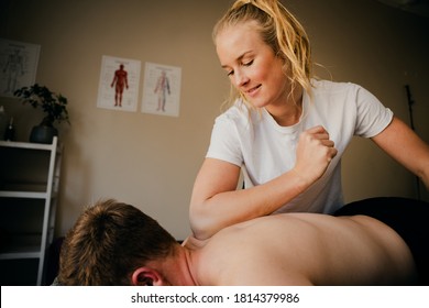 Young Blonde Caucasian Female Therapist Massaging With Elbow, Neck Of White Male Patient Lying On Bed In Studio.