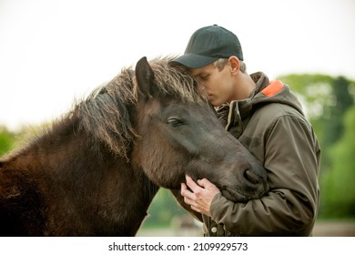 Young blonde caucasian man with cap petting a domesticated dark brown Icelandic mare. Human and animal equine friendship. 
