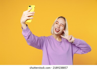 Young Blonde Caucasian Woman Bob Haircut Makeup In Basic Purple Shirt Close Up Doing Selfie Shot On Mobile Phone Blinking Showing Victory Sign With Tongue Isolated On Yellow Background Studio Portrait