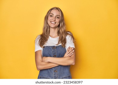 Young blonde Caucasian woman in denim overalls posing on a yellow background, who feels confident, crossing arms with determination.